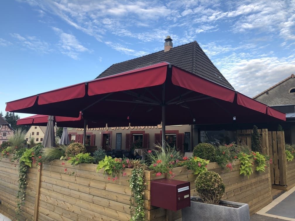 Un auvent rouge ombrage la terrasse d'un restaurant, devant laquelle se trouve un parterre de fleurs. Ciel bleu.