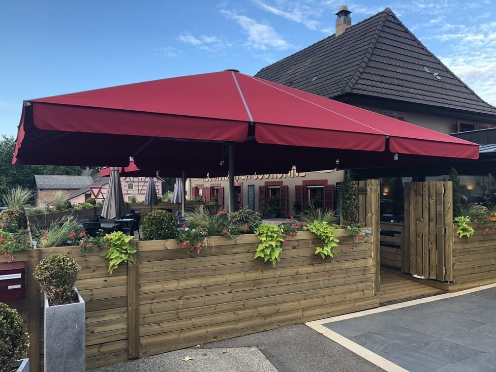 Un parasol rouge ombrage un coin salon extérieur avec une clôture en bois et des plantes en pot.
