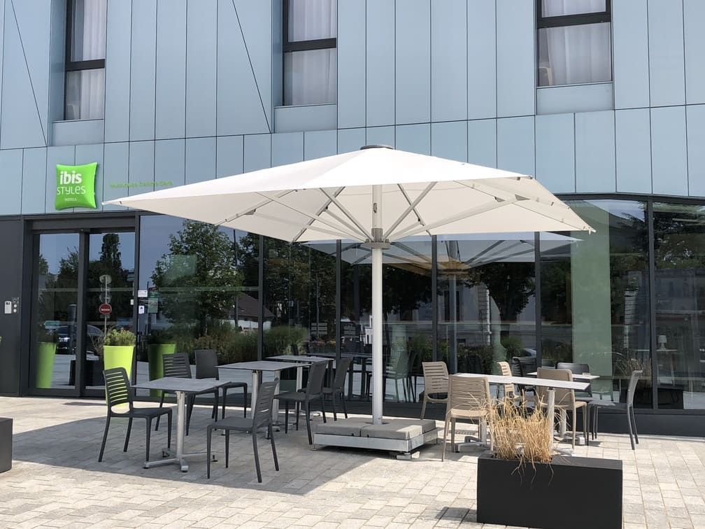 Terrasse extérieure avec tables et chaises sous un grand parasol blanc à l'entrée d'un hôtel Ibis Styles.
