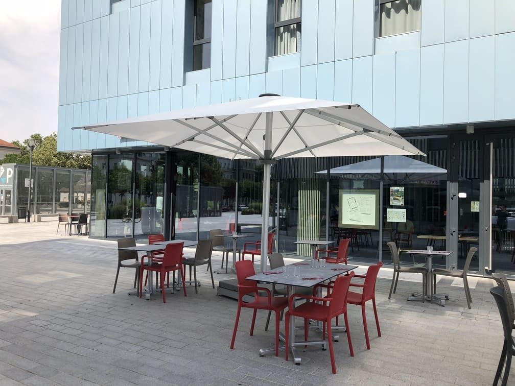 Terrasse de café avec chaises et tables rouges et grises, et un grand parasol blanc. Bâtiment moderne en arrière-plan.