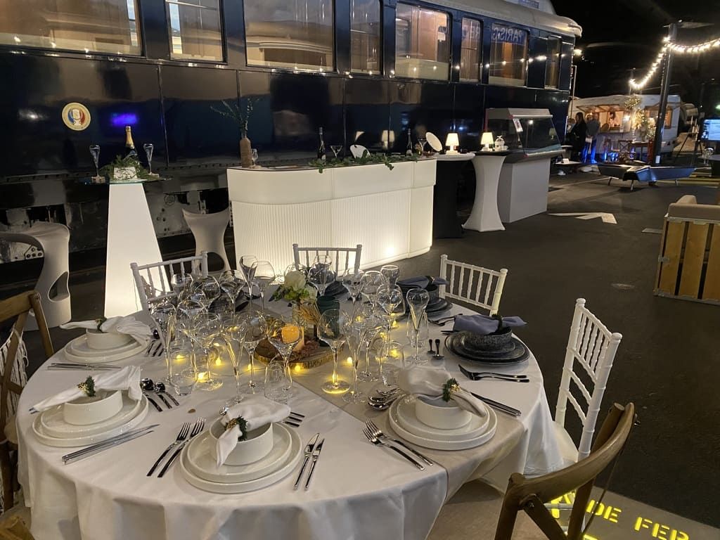 Une table ronde est dressée pour un dîner formel, avec un wagon de train en arrière-plan, éclairé d'une lumière tamisée.