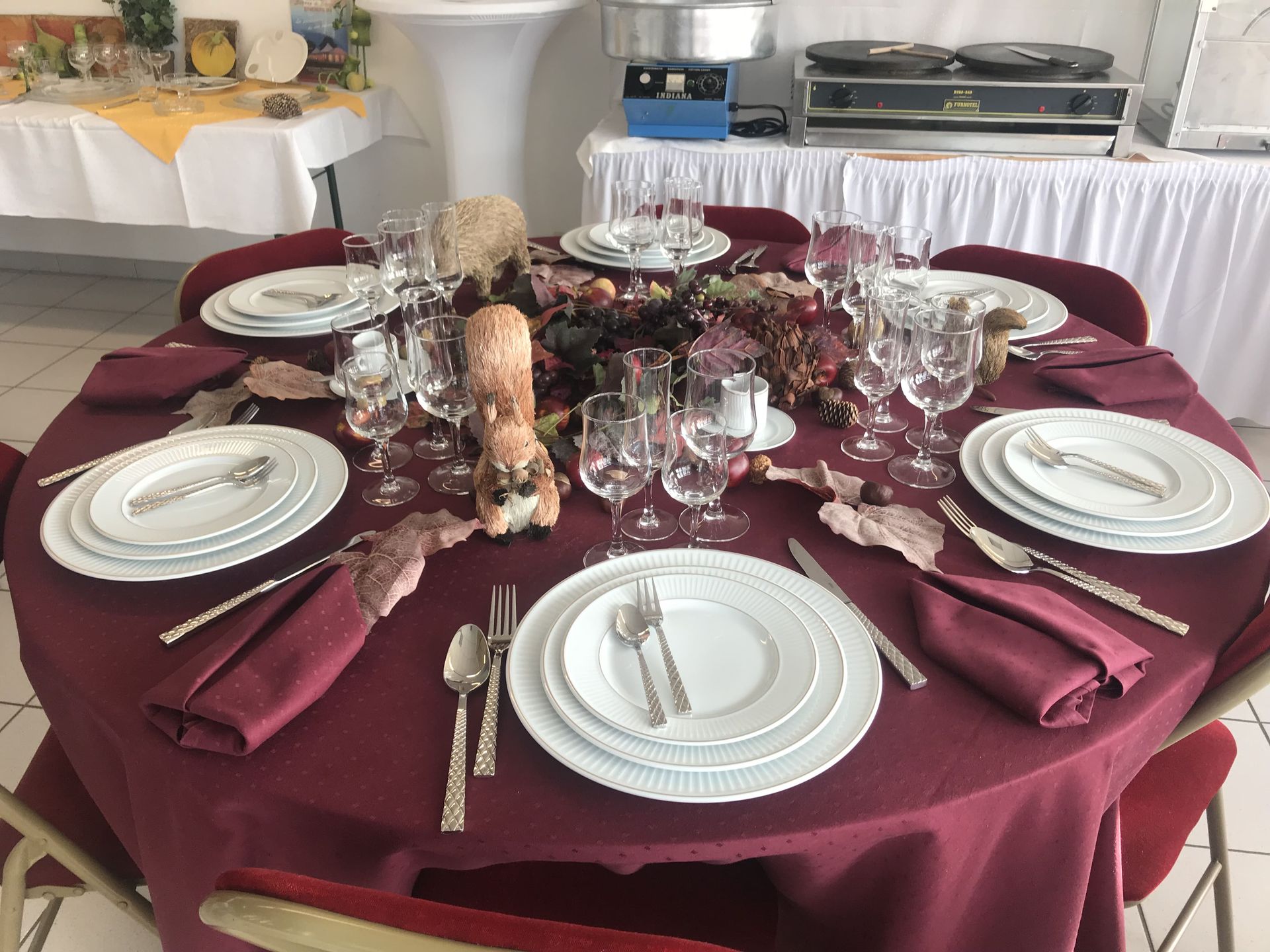 Une table ronde dressée pour un dîner formel, avec nappe bordeaux, assiettes, couverts et verres à vin.