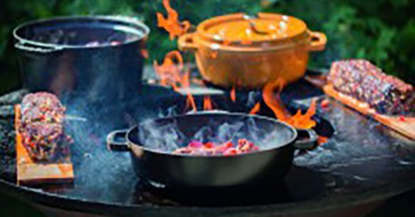 Cuisiner au feu de bois : marmites et viande sur des planches en bois, flammes, fumée.