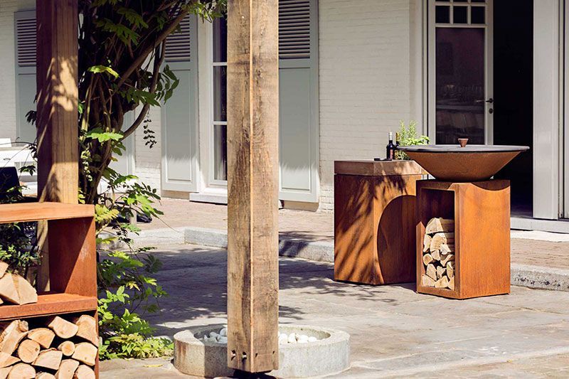 Terrasse extérieure avec un barbecue en métal rouillé posé sur un meuble de rangement en bois et en métal contenant du bois de chauffage.
