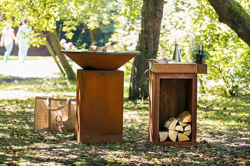 Foyer extérieur en métal rouillé et support à bois de chauffage dans un cadre évoquant un parc.