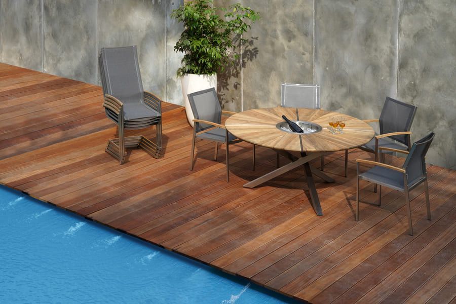 Ensemble de repas d'extérieur sur une terrasse en bois au bord d'une piscine. Table en teck avec chaises, une grande plante et un mur en béton.