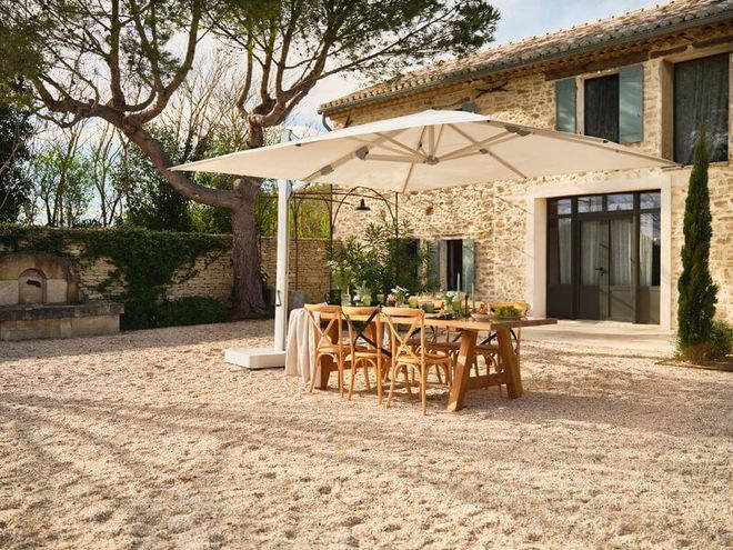 Terrasse aménagée sous un grand parasol dans une cour entourée de murs en pierre. Table dressée avec des chaises, à côté d'un bâtiment.