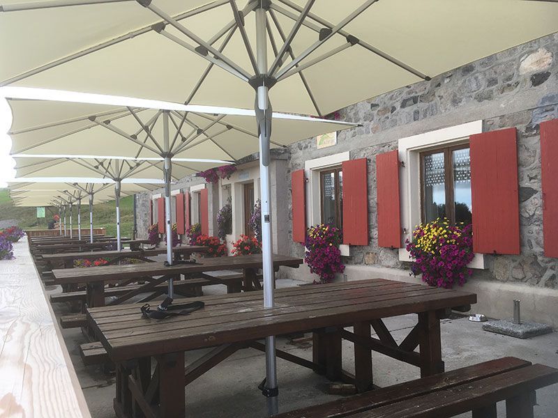 Café en plein air avec tables de pique-nique sous de grands parasols, à côté d'un bâtiment en pierre aux volets rouges.
