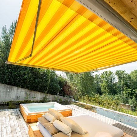 Auvent à rayures jaunes et orange au-dessus d'une terrasse en bois avec un jacuzzi et un coin salon extérieur.