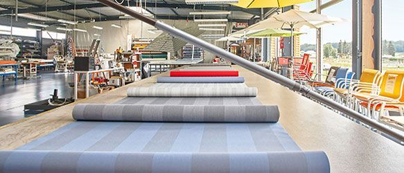 Des rouleaux de tissu rayé sont disposés sur une longue table dans un atelier, avec des sièges d'extérieur en arrière-plan.