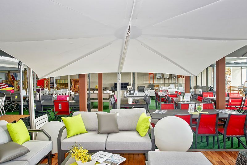 Magasin de mobilier d'extérieur avec parasol blanc. Canapés avec coussins verts et gris, tables et chaises.
