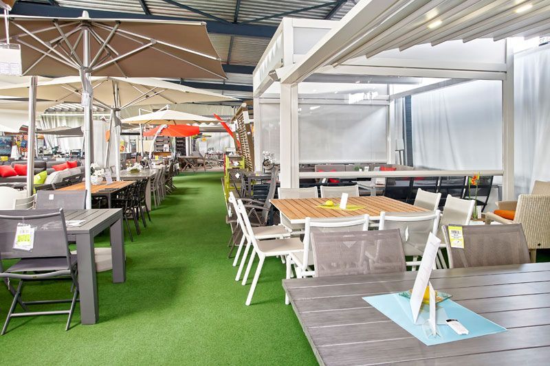 Salle d'exposition intérieure avec mobilier de terrasse : tables, chaises, parasols et pergola présentés sur une pelouse verte.