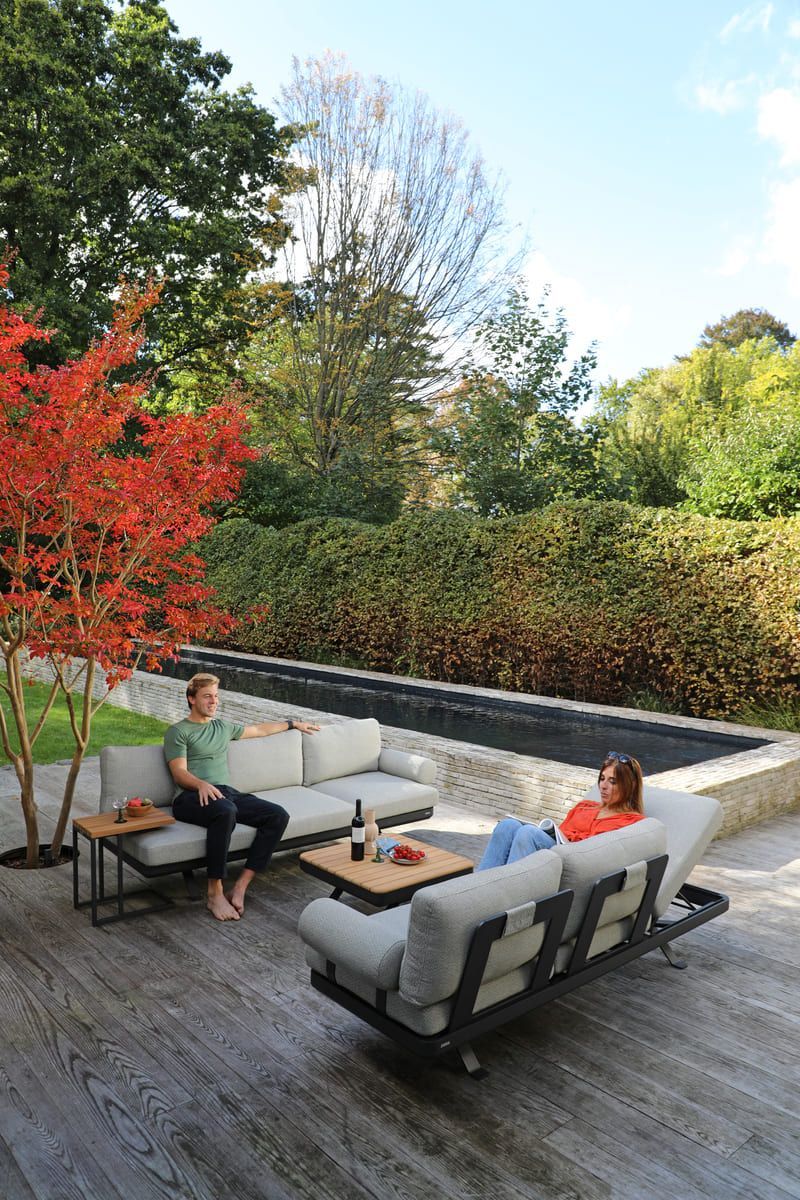 Deux personnes se détendent sur du mobilier de jardin moderne, sur une terrasse à côté d'une piscine sombre, sous des arbres aux couleurs d'automne.