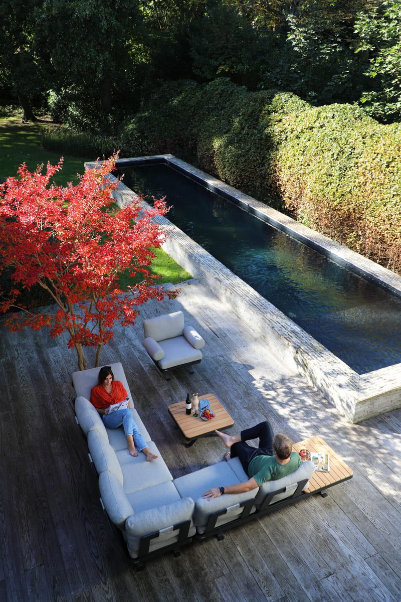 Terrasse en bois avec une longue piscine. Deux personnes se détendent sur un canapé d'angle gris, un arbre rouge à proximité.