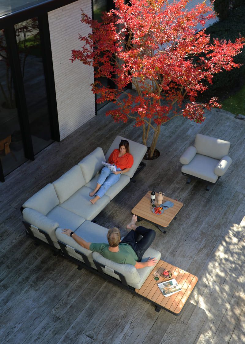Coin salon extérieur avec deux personnes se relaxant sur un canapé d'angle gris clair, de petites tables et un arbre aux feuilles rouges.
