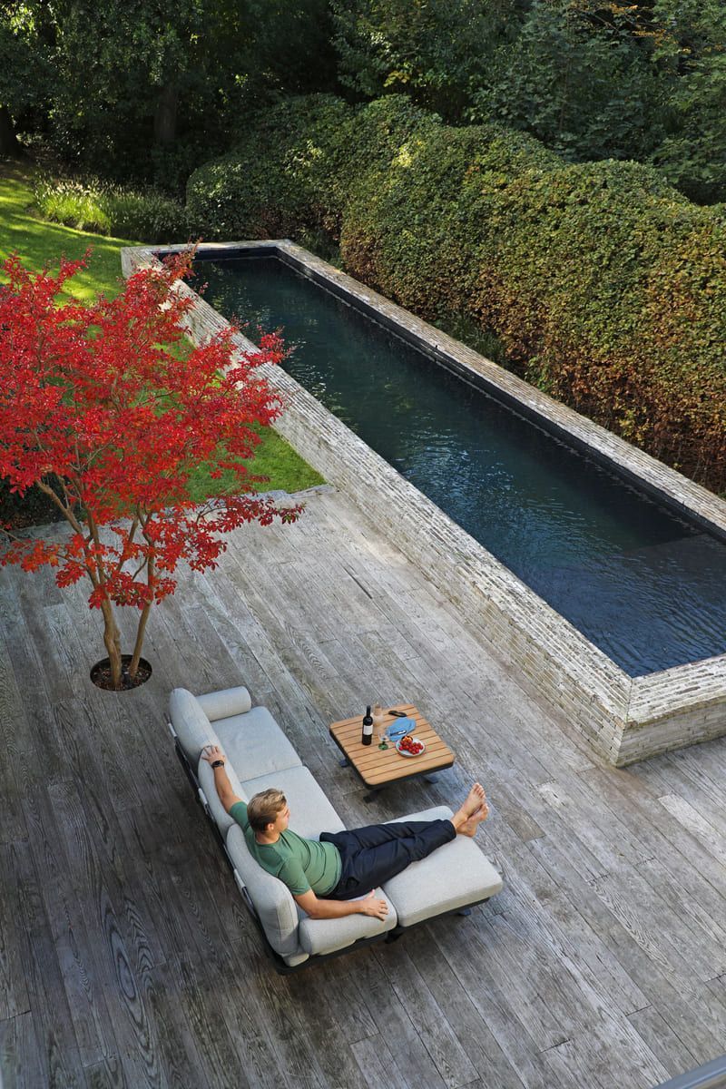 Un homme se détend sur une chaise longue à côté d'une longue piscine rectangulaire, d'un arbre rouge et d'une terrasse en bois.