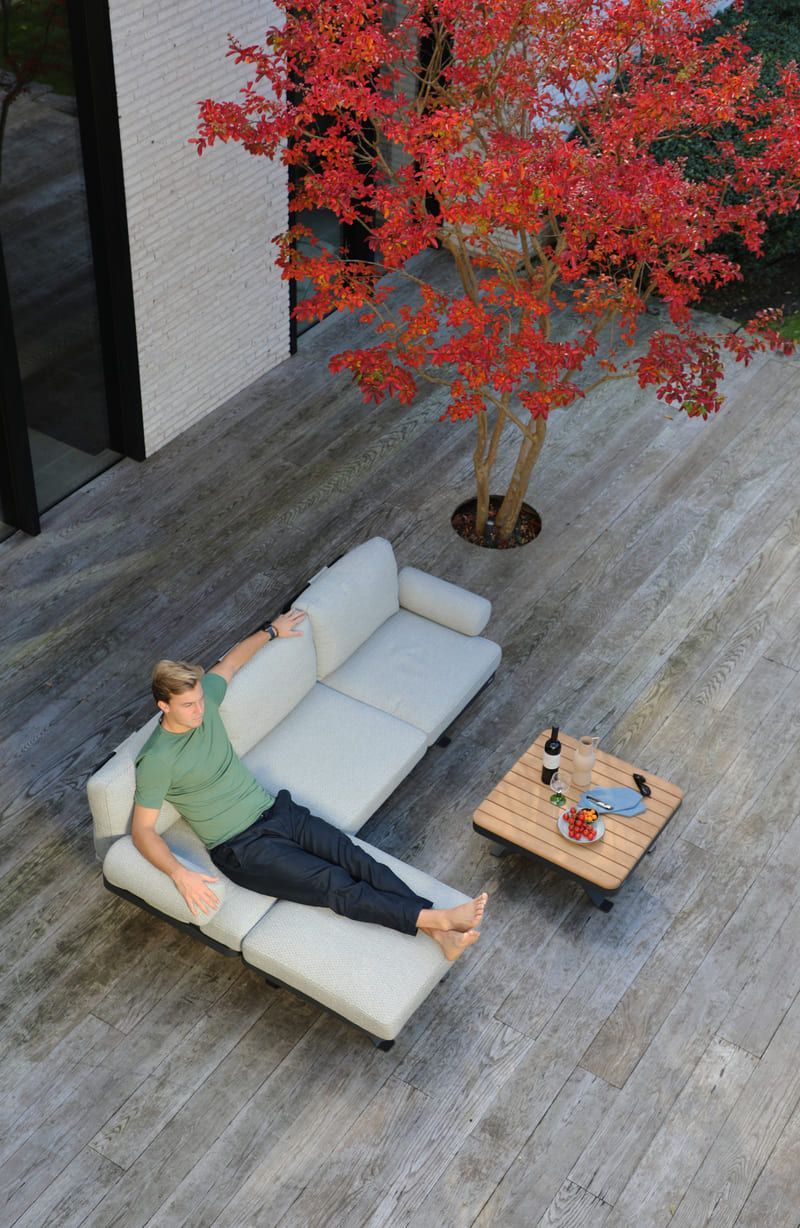 Un homme se détend sur un canapé d'angle beige d'extérieur, installé sur une terrasse avec une table. Un arbre rouge se profile à l'arrière-plan.