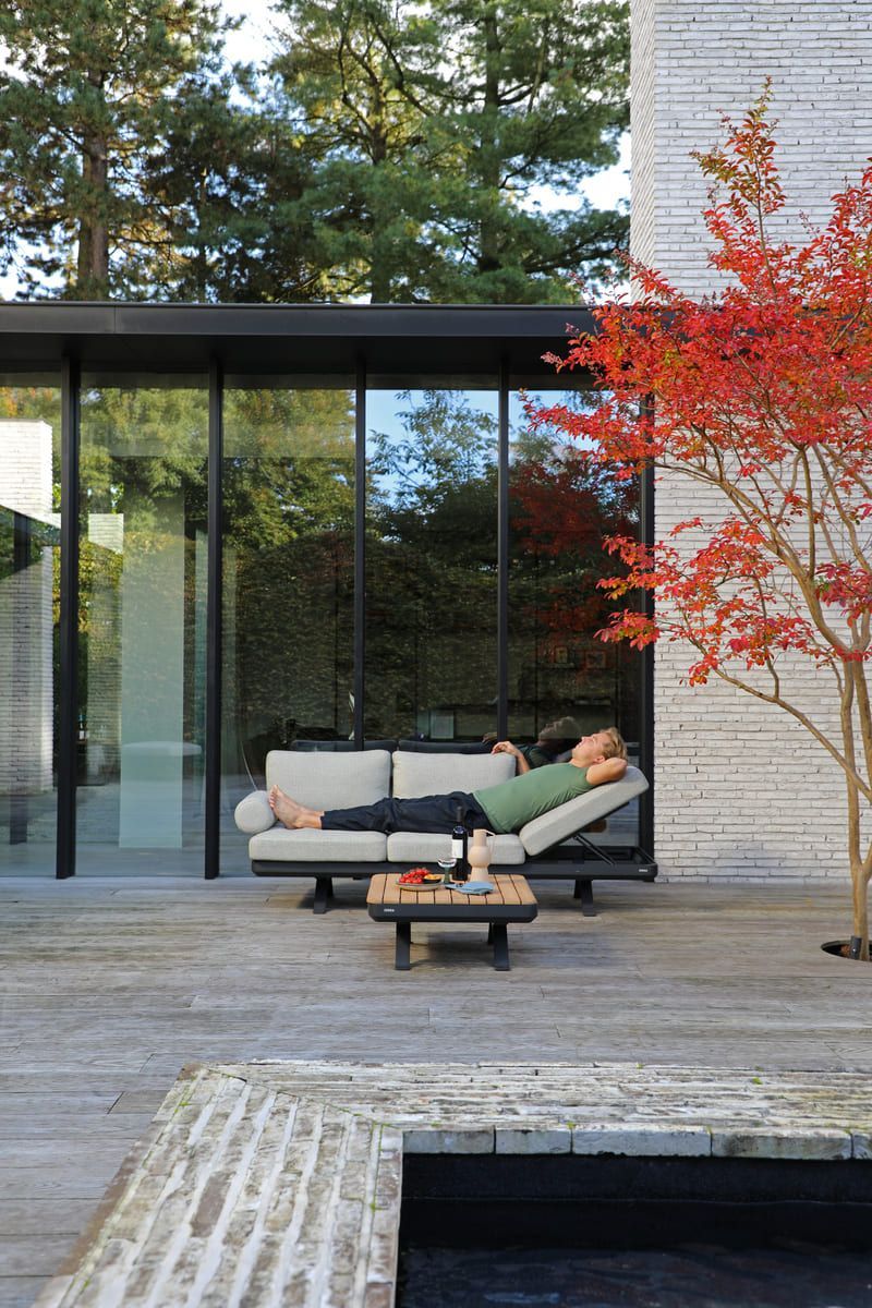 Une personne se détend sur un canapé d'extérieur à côté d'un arbre aux feuilles rouges et d'un bâtiment aux parois de verre.