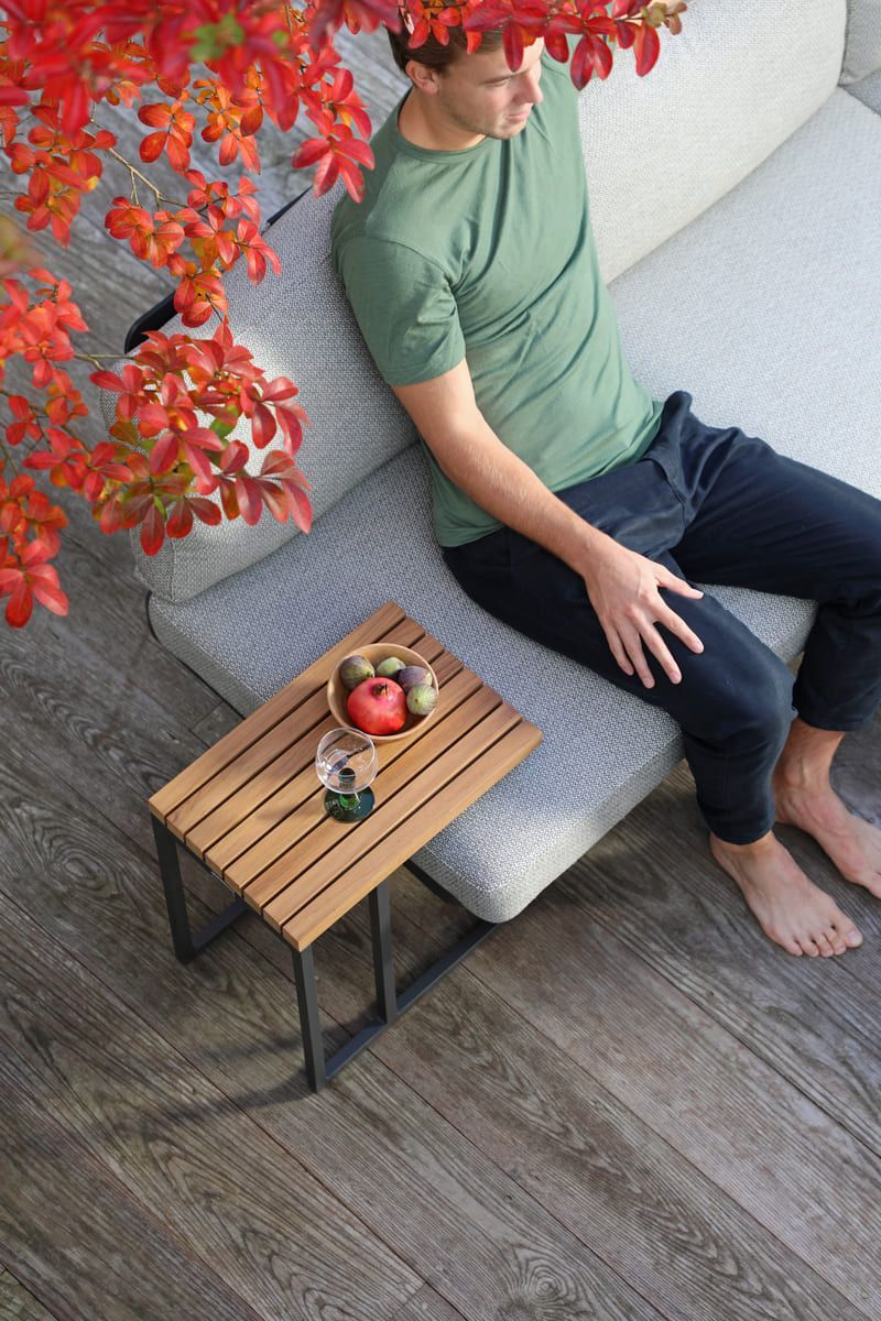 Un homme se détend sur un canapé d'extérieur, une petite table avec de la nourriture et des boissons, du feuillage rouge au-dessus de sa tête.
