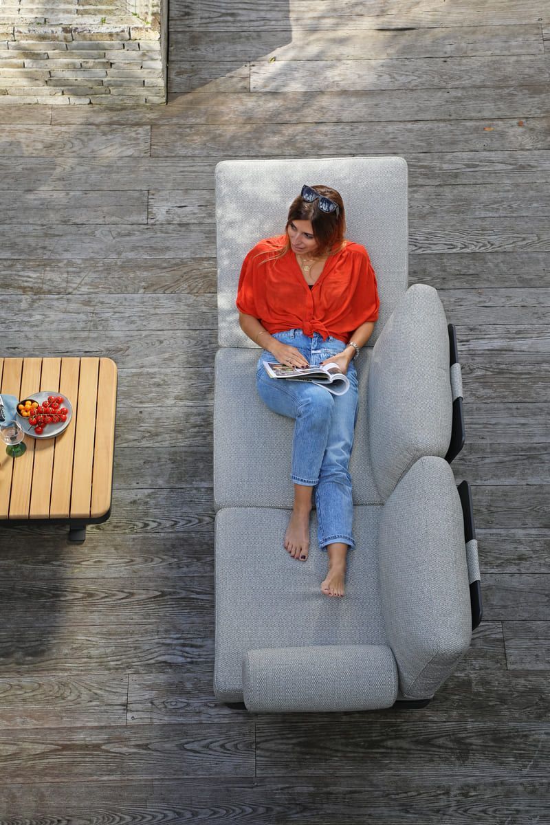 Femme en haut rouge et jean lisant sur une chaise longue grise sur une terrasse en bois.