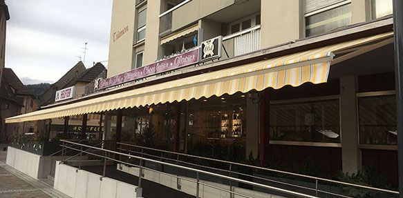 Façade d'un restaurant avec un auvent jaune. On aperçoit des bâtiments, une rampe d'accès et un ciel nuageux.