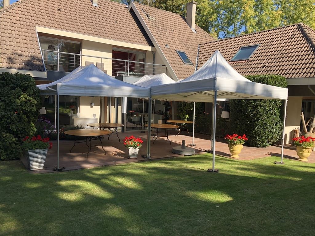 Terrasse aménagée sous une tente, avec tables et chaises installées sur un patio, une maison en arrière-plan.