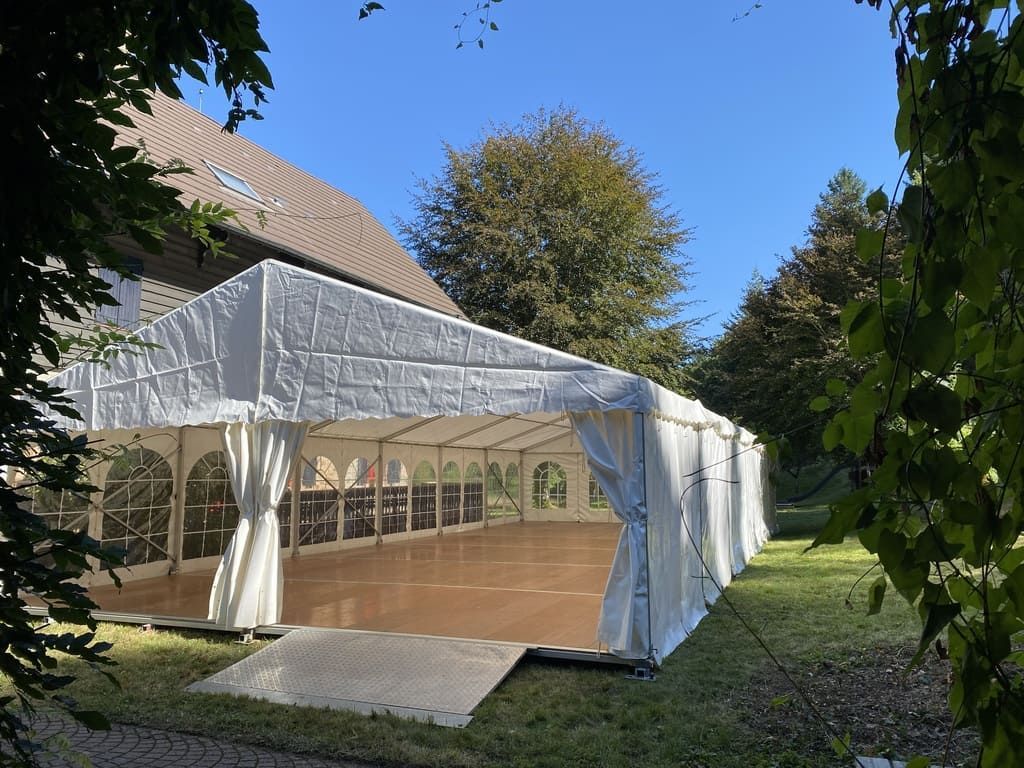 Une tente blanche, avec un plancher en bois, est installée sur une pelouse. En arrière-plan, on aperçoit un ciel bleu et des arbres.