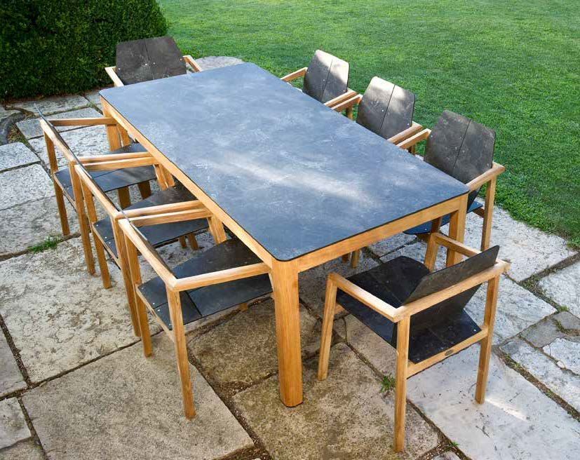 Table de salle à manger extérieure avec huit chaises sur une terrasse en pierre, devant une pelouse verte et des arbustes.