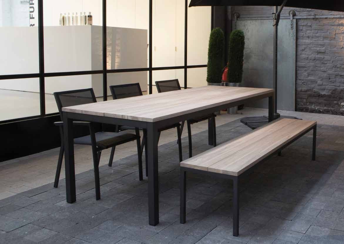 Ensemble de salle à manger d'extérieur : une table rectangulaire, un banc et des chaises sur une terrasse avec un mur de briques en arrière-plan.