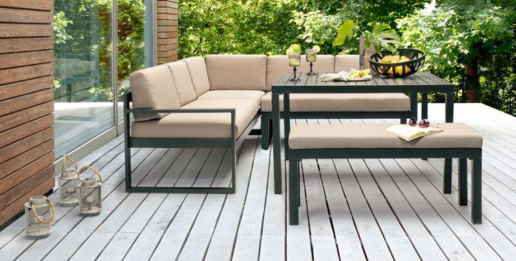 Ensemble table et canapé d'extérieur avec coussins beige clair sur une terrasse en bois, sur fond de verdure.
