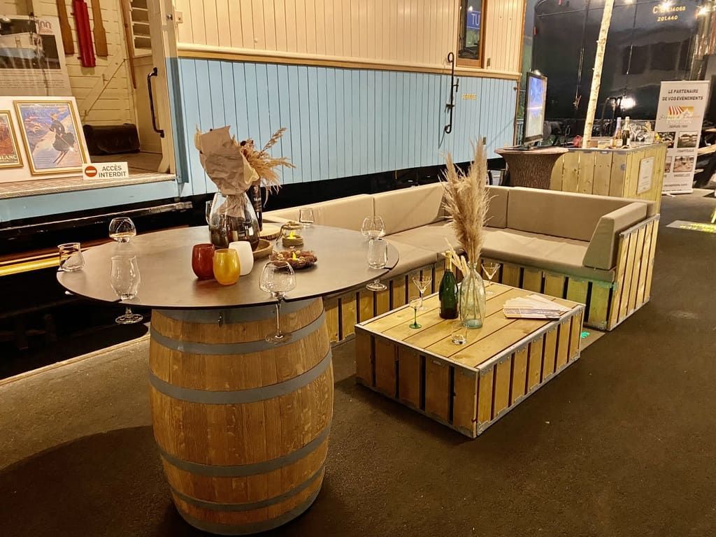 Coin salon extérieur avec table en forme de tonneau, canapé en caisse de bois, verres et plantes décoratives.