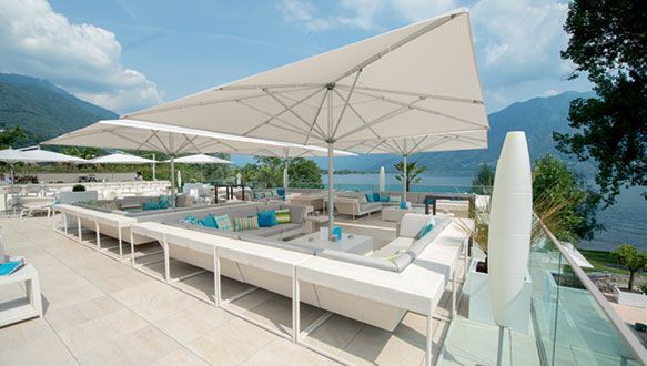 Salon extérieur avec canapés blancs, parasols et vue sur le lac sous un ciel bleu.
