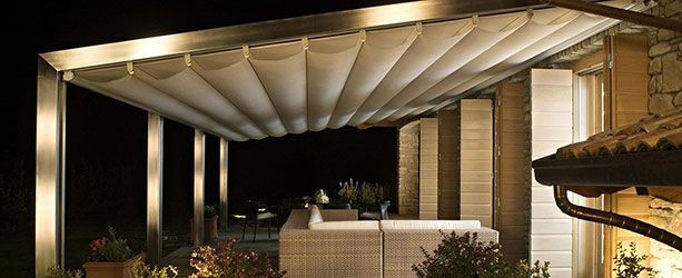 Terrasse extérieure avec auvent rétractable la nuit. On aperçoit des colonnes éclairées, un canapé et des plantes.