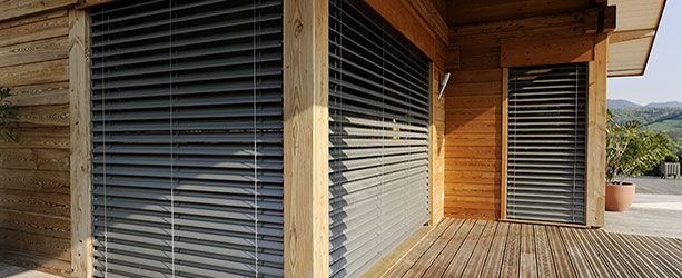 Bâtiment en bois avec stores horizontaux gris fermés. Vue extérieure avec terrasse et végétation en arrière-plan.
