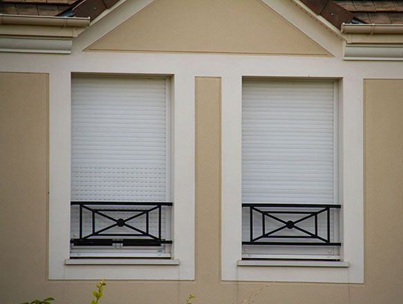Deux fenêtres à volets blancs et balustrades décoratives noires sur un bâtiment beige.