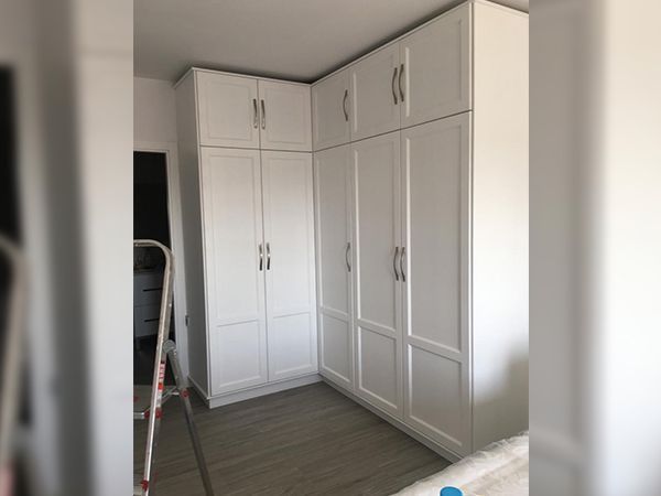 Armario de esquina blanco con puertas con paneles elevados y tiradores plateados en una habitación con suelo gris.