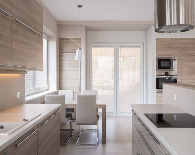Cocina moderna con gabinetes de madera clara, encimeras blancas y mesa de comedor. Grandes ventanales que aportan luz natural.