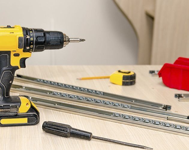 Taladro amarillo y negro, destornillador, correderas para cajones y cinta métrica sobre una superficie de madera clara; herramientas para montaje de muebles.