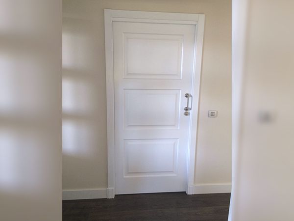 Puerta con paneles blancos en un pasillo beige. Suelo oscuro visible bajo la puerta.