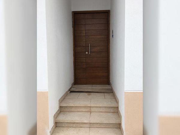 Una puerta de madera con un largo tirador de plata, situada al final de un estrecho pasillo con escalones de baldosas y paredes blancas.