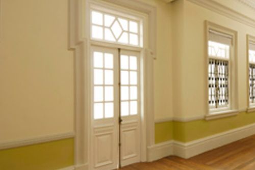 Un pasillo interior con puertas y ventanas dobles, todas con marcos blancos. Las paredes son de color amarillo claro con un zócalo verde.