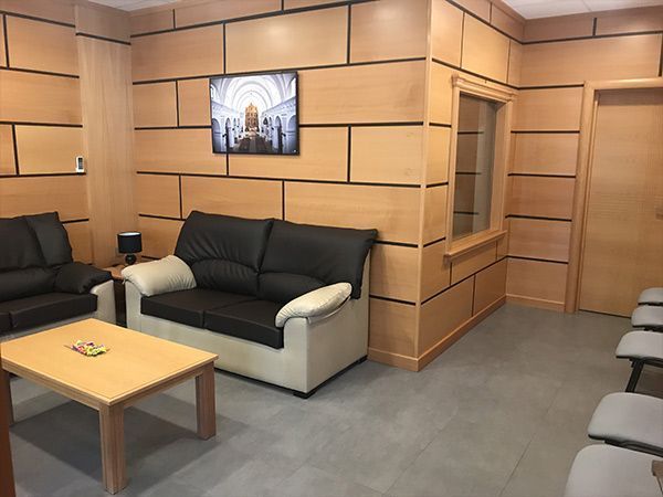 Una sala de espera con paneles de madera clara, suelo de baldosas grises, sofás de cuero negro, una mesa de centro y algunas sillas. Un cuadro enmarcado cuelga de la pared.