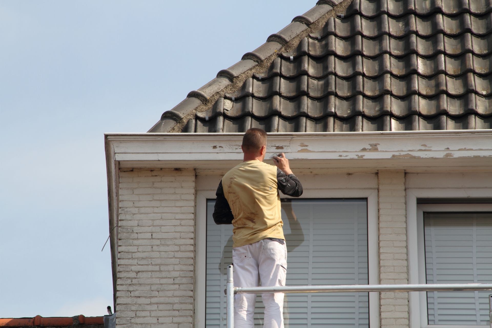 Une personne sur un échafaudage peint l'avant-toit d'un bâtiment en briques blanches sous un toit de tuiles. Lien vers la page enduit projeté.