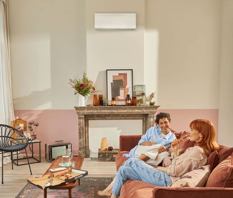 Deux jeunes femmes dans un salon beige et rouge pastel avec une fausse cheminée surmontée d'une climatisation