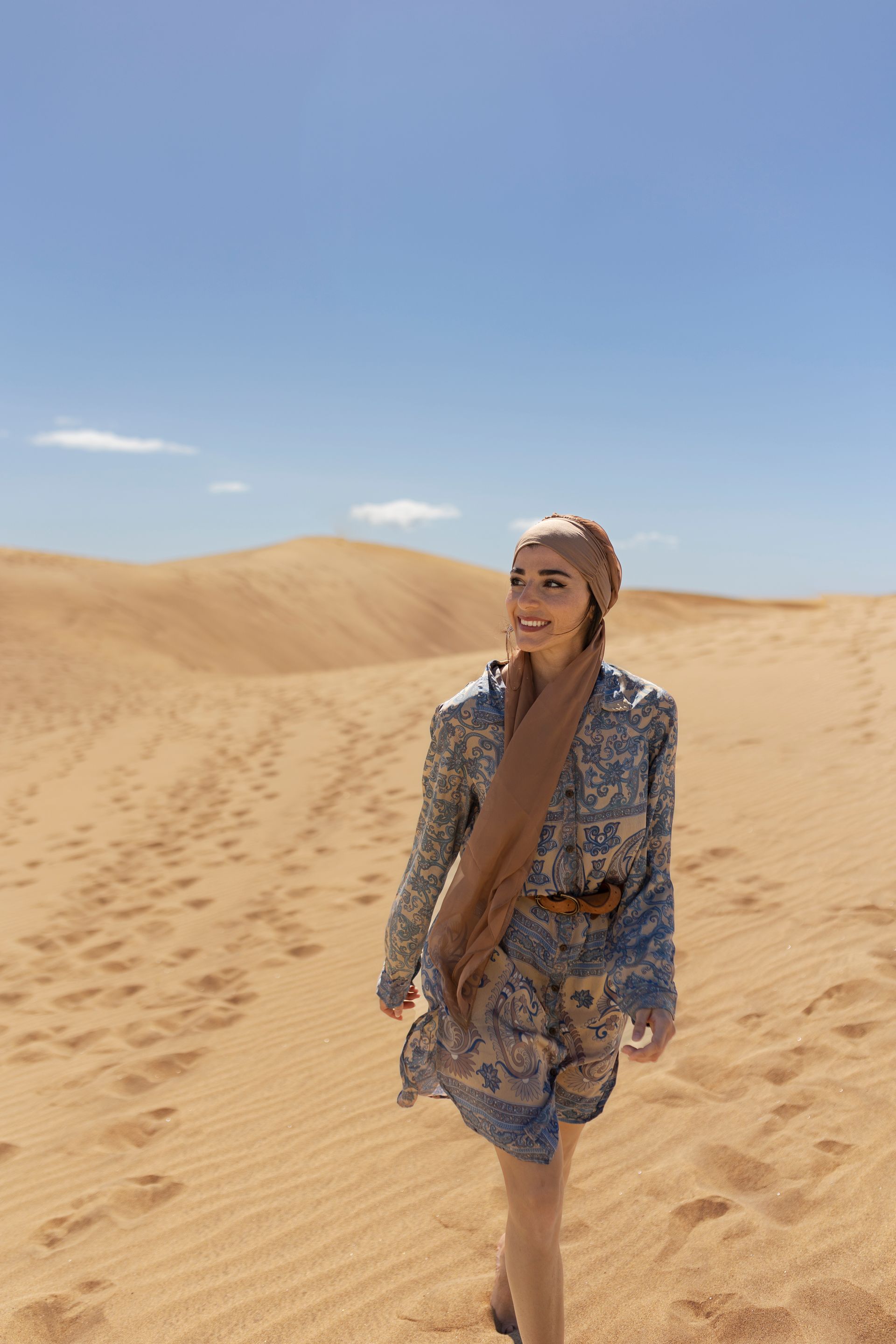 Une femme en robe à motifs et foulard sur la tête marche sur des dunes désertiques ensoleillées sous un ciel bleu.