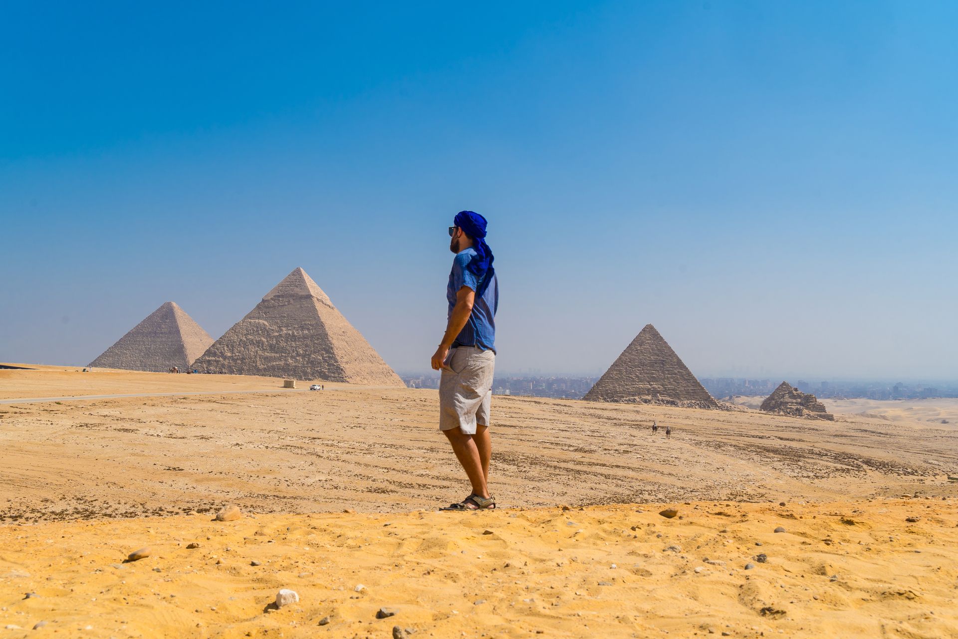 Une personne se tient debout dans le désert, avec les pyramides en arrière-plan, sous un ciel bleu clair.