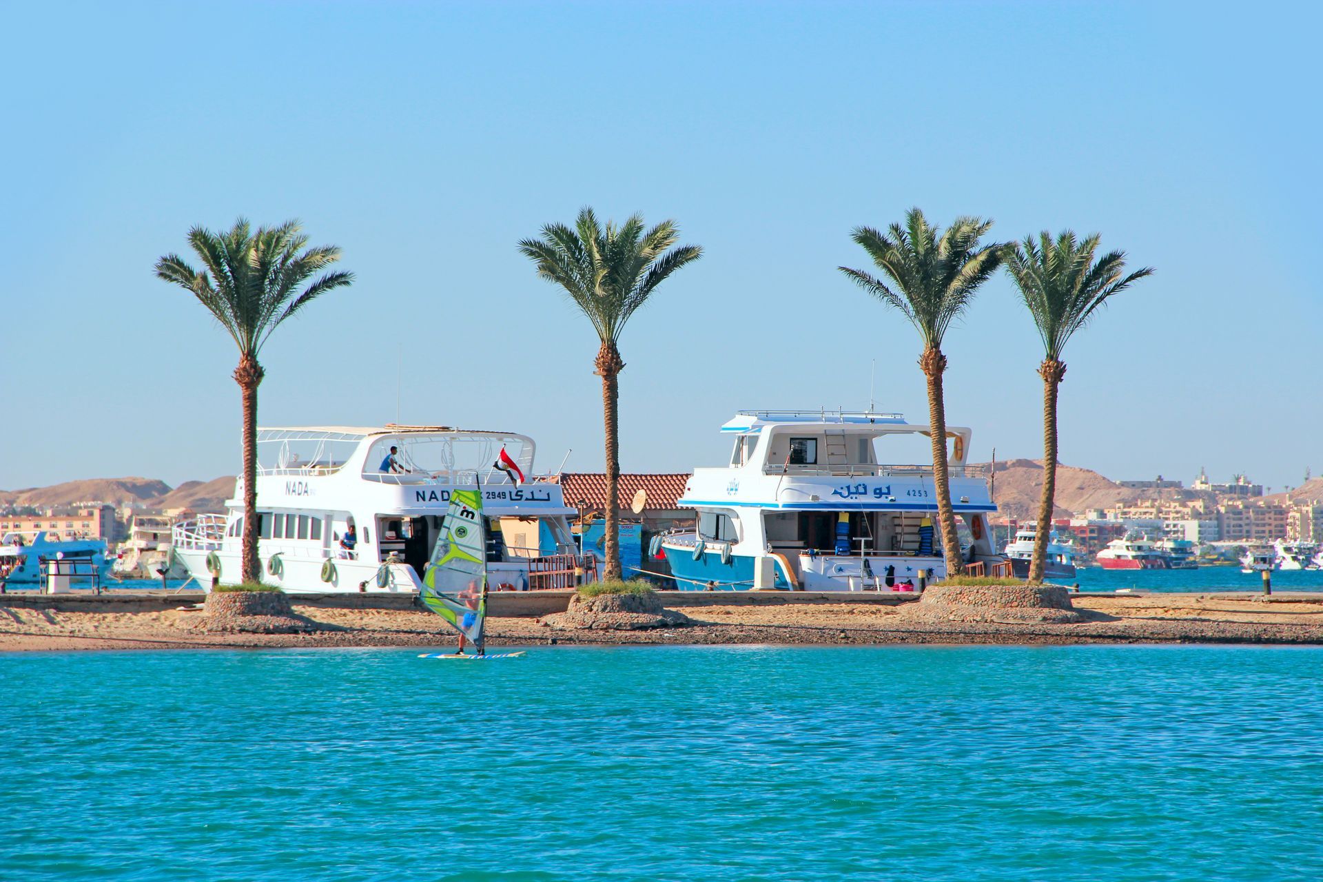 Des yachts blancs et des palmiers sur une petite île, avec une eau turquoise au premier plan et la silhouette d'une ville au loin.