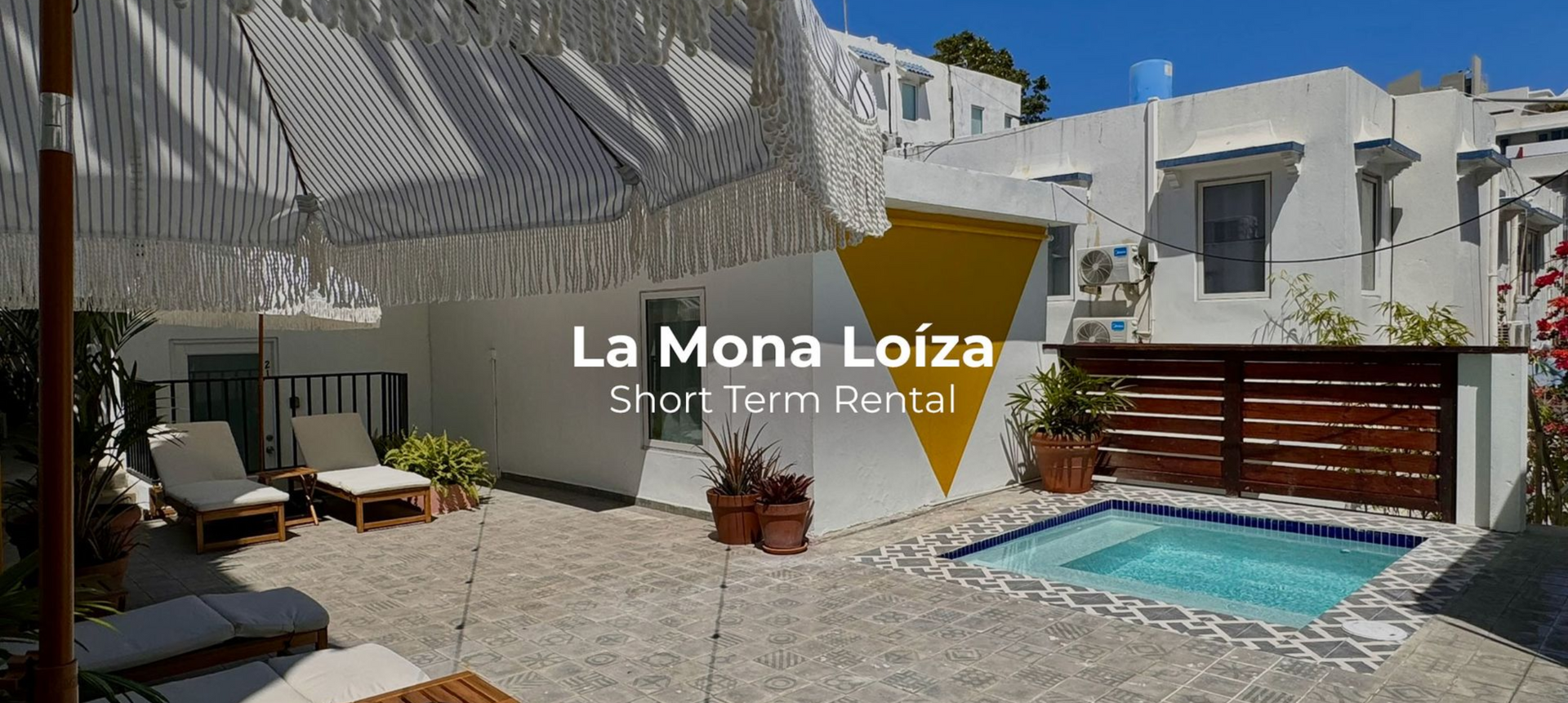 Outdoor patio with small plunge pool, white lounge chairs under a striped fringe umbrella, white buildings with a bold yellow triangle wall design, potted plants, and clear blue sky in the background.