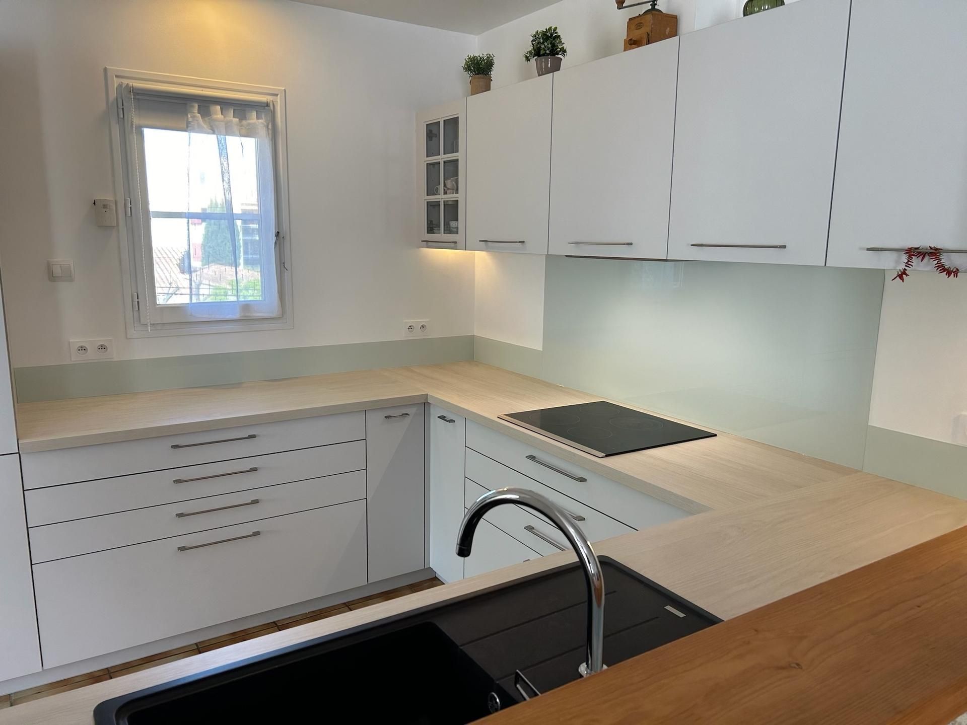 Cuisine blanche avec comptoirs en bois clair, évier noir et plaque de cuisson intégrée. Petite fenêtre.