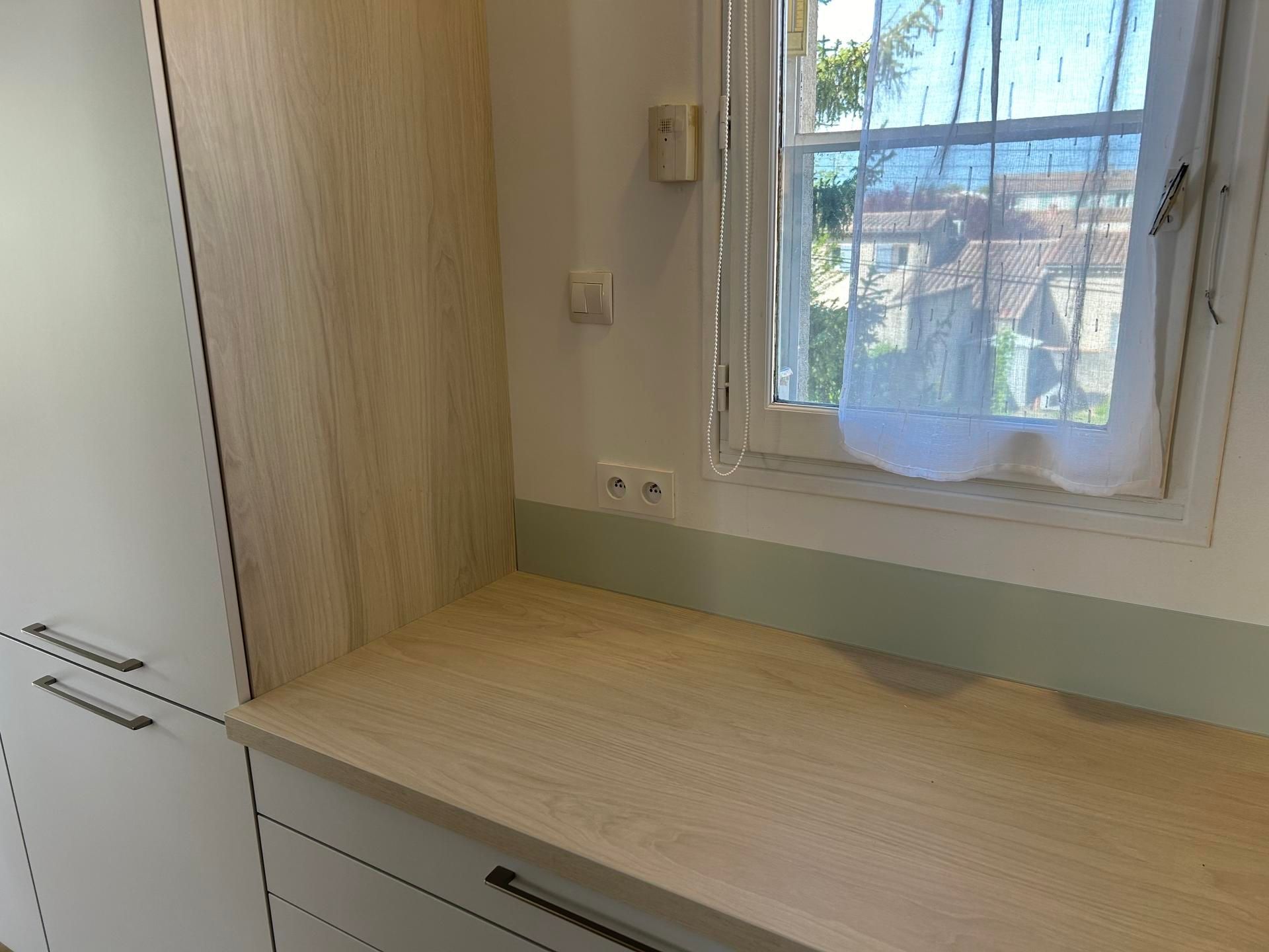 Comptoir de cuisine en coin avec fenêtre, armoires en bois clair et murs blancs.